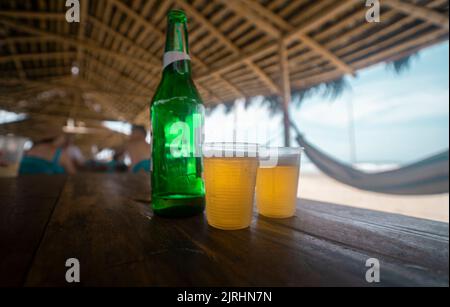 Deux verres en plastique avec bière et une bouteille verte sur une table en bois dans un restaurant de plage avec un toit en bambou et des hamacs sur les côtés pendant un d ensoleillé Banque D'Images