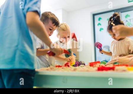 Détente sensorielle jeu avec sable cinétique moulable à l'école maternelle. Les tout-petits avec leur professeur s'amusent autour de la table en utilisant différents outils pour sculpter le sable, tels que des punaises texturées et colorées, des pinces coupantes, des étuis en silicone. Compétences motrices fines et développement créatif . Photo de haute qualité Banque D'Images