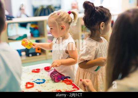 Détente sensorielle jeu avec sable cinétique moulable à l'école maternelle. Les tout-petits avec leur professeur s'amusent autour de la table en utilisant différents outils pour sculpter le sable, tels que des punaises texturées et colorées, des pinces coupantes, des étuis en silicone. Compétences motrices fines et développement créatif . Photo de haute qualité Banque D'Images