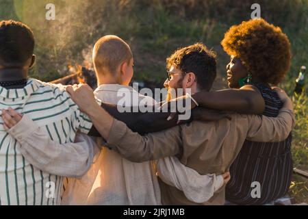 Vue arrière de multiculturel bon vieux amis qui embrasse et d'avoir un pique-nique de fin de semaine dans la nature près du feu de camp, parler et rire. Heure du coucher du soleil. Photo de haute qualité Banque D'Images