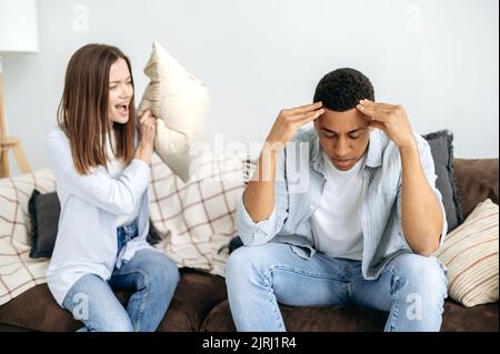 Un couple multiracial marié règle les choses tout en étant assis dans le salon sur un canapé, une femme caucasienne crie à son mari hispanique. Malentendu dans les relations, jalousie, querelle Banque D'Images