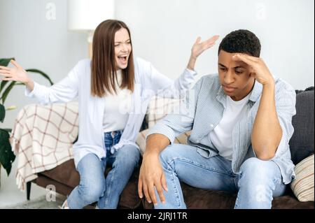 Malentendu dans les relations, jalousie, querelle. Un couple multiracial marié règle les choses tout en étant assis dans le salon sur un canapé, une femme caucasienne crie à son mari hispanique Banque D'Images