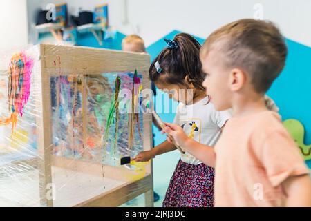 Peinture de film adhésif. Peinture pour tout-petit avec une éponge, des pinceaux et des peintures sur un film adhésif enroulé autour de l'étagère en bois. Un enseignant les aidant. Activité créative pour les enfants compétences sensorielles, l'attention spatiale et le développement perceptuel visuel à l'école maternelle. Photo de haute qualité Banque D'Images