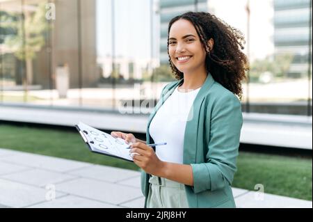 Portrait d'une femme d'affaires intelligente réussie, de nationalité brésilienne ou hispanique, consultant financier, mentor, debout à l'extérieur, tenant des documents financiers dans sa main, regardant la caméra, souriant Banque D'Images