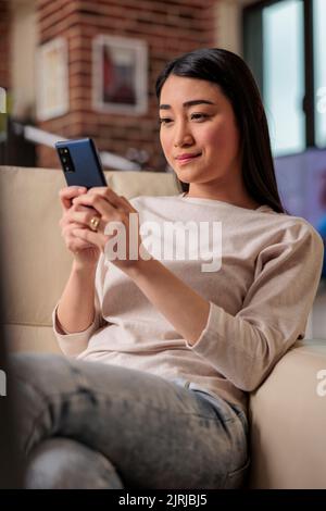 Femme asiatique utilisatrice de smartphone à la maison sur un canapé, regardant le téléphone avec un sourire authentique. Utilisateur mobile de la technologie Web Internet Banque D'Images