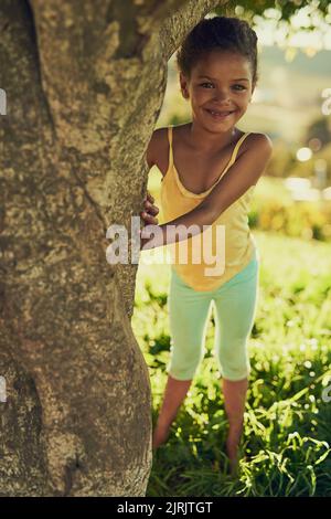 Personne ne me trouvera ici. Portrait d'une fille souriante jouant dehors derrière un arbre. Banque D'Images