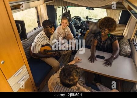 Joyeux groupe interracial de bons amis sur une route d'été, assis à l'intérieur de leur confortable motorhome moderne, jouer de la guitare, parler et rire. Intérieur du RVS. Photo de haute qualité Banque D'Images