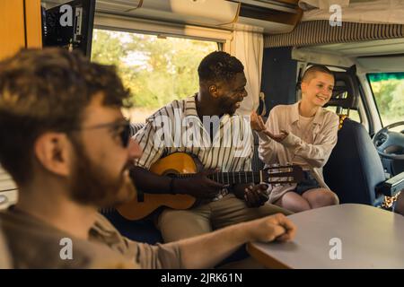 Groupe multi-ethnique d'amis proches lors d'un voyage en voiture d'été dans un camping-car, assis dans leur confortable et spacieux motorhome, jouer de la guitare, parler et rire de coeur. Photo de haute qualité Banque D'Images