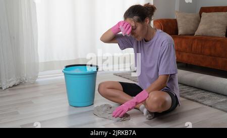 Une femme fatiguée en gants de caoutchouc lave le sol avec un chiffon sur ses genoux Banque D'Images