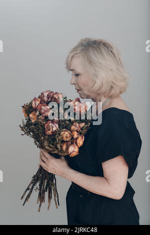 Triste femme âgée senior en chemise blanche tient avec une vieille fleur de rose mort sèche. Notion de vieillesse, de vieillissement et de mort. Banque D'Images