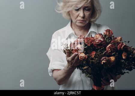 Triste femme âgée touchant un bouquet de fleurs de rose flétrisées. Femme adulte aux cheveux gris dans la dépression. Concept de vieillissement ancien. Studio Shoot isolé sur fond gris. Copier l'espace Banque D'Images