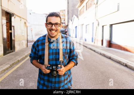 Joyeux jeune homme charismatique photographe dans des vêtements décontractés debout près du bâtiment de merde et regardant l'appareil photo Banque D'Images