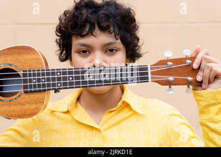 Femme ethnique confiante en chemise jaune tenant une guitare ukulele face regardant la caméra Banque D'Images