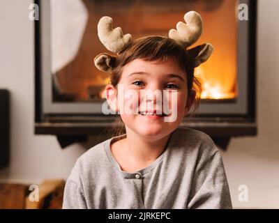 Une fille stupéfait dans un bandeau de rennes regardant l'appareil photo tout en étant assise dans le salon pendant la fête de Noël Banque D'Images