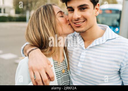 Petite amie à contenu jeune qui embrasse la joue d'un ami heureux debout dans la rue de la ville et embrassant avec amour Banque D'Images