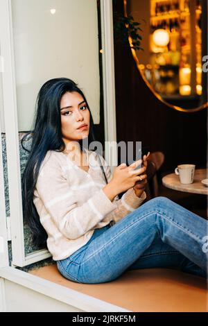 Femme ethnique avec téléphone portable regardant la caméra tout en étant assis sur le rebord de fenêtre de la cafétéria Banque D'Images