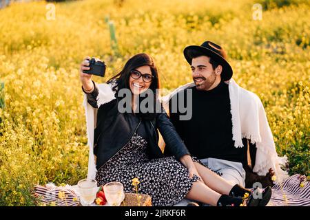 Jeune femme ethnique gaie avec un petit ami qui prend son propre portrait sur un appareil photo tout en étant assise sur un pré Banque D'Images