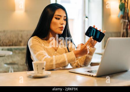 Jeune femme ethnique montrant un écran noir sur un téléphone portable tout en parlant pendant le chat vidéo sur netbook à table de café Banque D'Images
