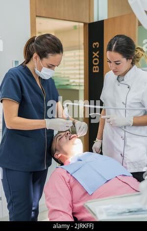 Femme en uniforme médical utilisant un outil professionnel sur les dents d'un homme adulte avec une infirmière assistante travaillant dans une clinique dentaire contemporaine Banque D'Images
