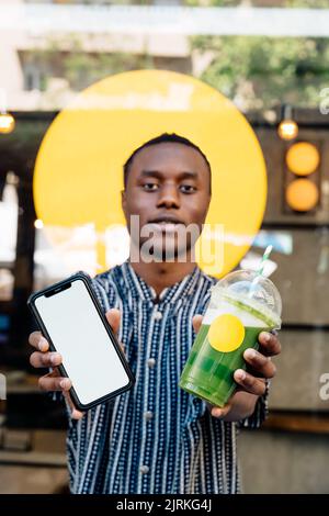 Contenu jeune homme noir faisant la démonstration d'un téléphone portable avec écran vierge et verre jetable de boisson saine tout en regardant l'appareil photo à l'extérieur Banque D'Images