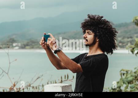 Vue latérale d'un homme afro-américain prenant des photos de l'océan sur un téléphone portable tout en se tenant sur un remblai urbain Banque D'Images