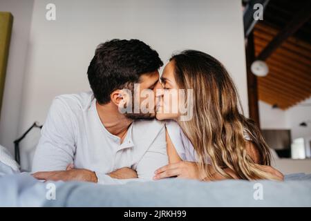 Jeune couple portant des vêtements de nuit similaires allongé sur un lit confortable avec des baisers de literie blanche Banque D'Images