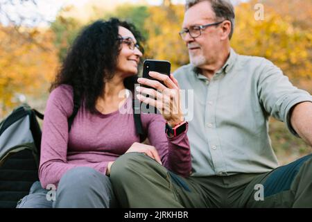 Content couple senior en vêtements décontractés et lunettes de vue parcourant un smartphone moderne tout en étant assis sur un sol herbeux dans le parc d'automne Banque D'Images