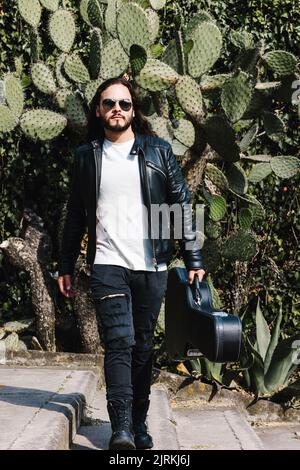 Guitariste latino-américain tendance avec veste en cuir et lunettes de soleil et étui de transport avec guitare, tout en regardant loin sur l'escalier contre les feuilles de cactus Banque D'Images