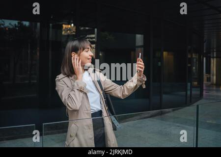 Jeune femme positive dans les écouteurs ayant chat vidéo sur smartphone près du bâtiment moderne dans la rue de la ville Banque D'Images