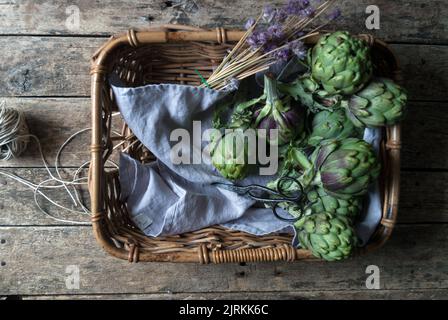 Vue d'en haut des artichauts verts frais posés dans un panier en osier sur une serviette décorée de fleurs sur fond de bois Banque D'Images