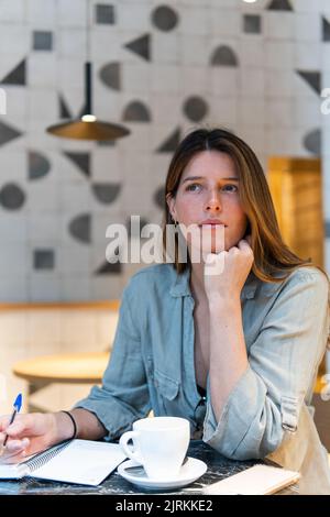 Jeune femme agité se penchée à la main à la table avec une tasse de boisson chaude et un bloc-notes tout en regardant loin dans la cafétéria Banque D'Images