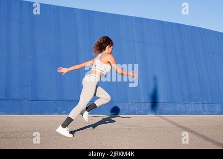 Vue latérale de la coureuse ciblée dans des vêtements de sport qui se rince sur la chaussée avec des nuances contre le mur bleu pendant l'entraînement Banque D'Images