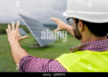 Homme en casque et gilet gesticalant et planifiant la construction de panneaux photovoltaïques contre ciel nuageux sur la station solaire Banque D'Images