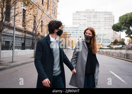 Joyeux jeune homme et femme dans des masques protecteurs tenant les mains et parlant amical en traversant la rue urbaine dans la ville d'automne Banque D'Images