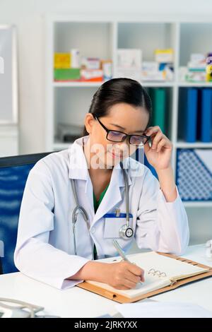 Souriant, un joli médecin asiatique prend des notes dans son planificateur Banque D'Images