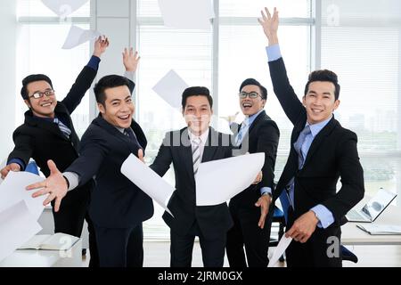 Des managers asiatiques heureux portant des costumes regardant l'appareil photo avec des sourires crasseux et jetant des documents dans l'air après avoir réussi le projet prometteur, portrait de groupe Banque D'Images