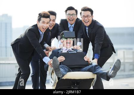 Portrait de groupe d'hommes d'affaires asiatiques joyeux portant des costumes élégants posant pour la photographie sur la grande terrasse du bâtiment de bureau et poussant la chaise pivotante avec leur collègue portant un casque VR Banque D'Images