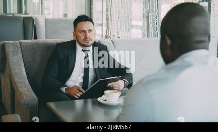 Portrait d'un homme d'affaires caucasien barbu vêché de vêtements formels prenant un entretien avec son employé américain Africain dans un café glassy pendant la pause déjeuner. Tasse à café et bloc-notes sont sur sa table Banque D'Images