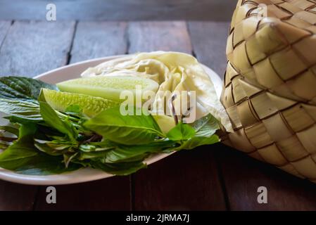 Panier de riz collant en osier de bambou appelé 'Kratip' dans la province du nord-est, en Thaïlande, servi avec un plat d'accompagnement de légumes sur la table en bois. Banque D'Images