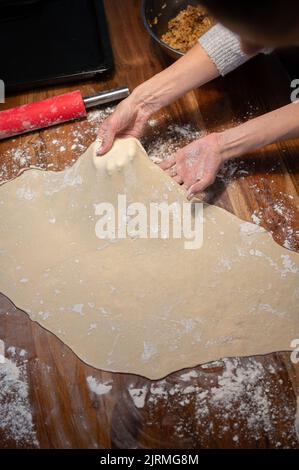 Vue depuis le dessus d'une femme tirant et étirant la pâte à pâtisserie maison pour strudel ou tarte sur une table à manger domestique de farine saupoudrée. Banque D'Images