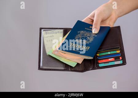 Passeport brésilien dans une main avec portefeuille, cartes de crédit et billets en euros sur fond blanc. Banque D'Images