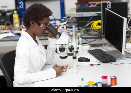Scientifique afro-américaine femme travaillant en laboratoire avec des instruments de technologie électronique et un microscope. Recherche et développement d'appareils électroniques par la femme noire. Banque D'Images