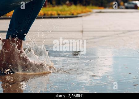 Femme portant des bottes de caoutchouc de pluie marchant courir et sautant dans le flaque avec des éclaboussures d'eau et des gouttes dans la pluie d'automne. Banque D'Images