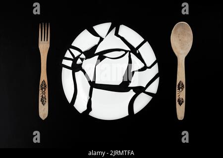 Plaque blanche cassée reconstruite sur une forme ronde isolée sur fond noir, avec une fourchette et une cuillère en bois. Art mural abstrait de cuisine Banque D'Images