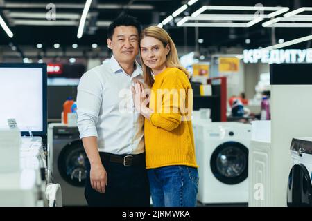 Portrait d'un jeune couple marié magasiner dans un supermarché d'appareils ménagers, une famille diversifiée d'un homme et d'une femme souriant et regardant l'appareil photo, le choix et l'achat d'appareils électroménagers Banque D'Images