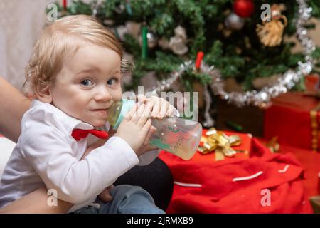 Un petit garçon boit dans une bouteille près d'un arbre de Noël avec des cadeaux Banque D'Images