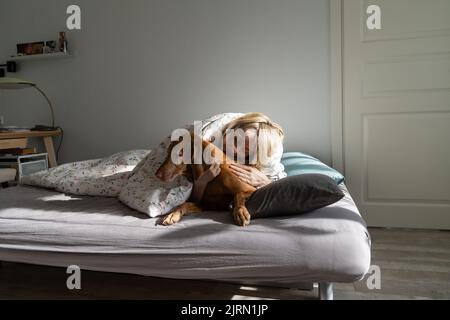 Une femme d'âge moyen se trouve avec un chien préféré recouvert d'une couverture chaude sur un lit double Banque D'Images