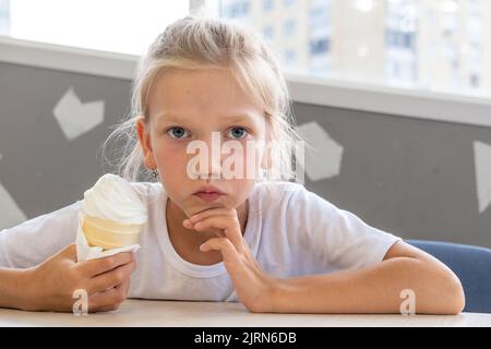 Portrait d'une petite fille drôle et affamée qui mange de la glace froide délicieuse dans une tasse de gaufres assis dans un café. Jour d'été. Un enfant aime la glace. Un suédois Banque D'Images