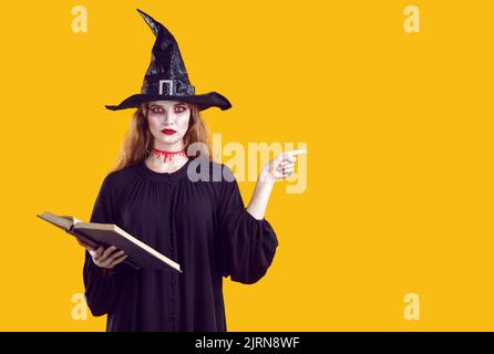 Femme créepy sérieuse dans le personnage de sorcière pointant du doigt sur le côté sur fond orange en studio. Banque D'Images