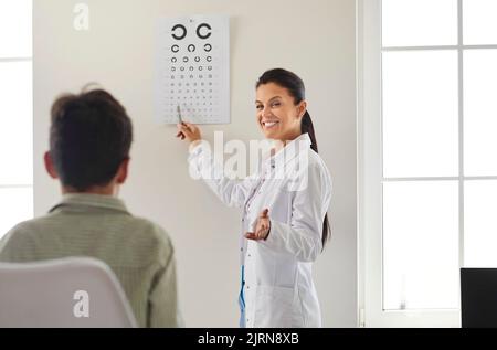 Un ophtalmologiste heureux qui teste la vue de l'enfant, qui pointe vers le graphique des yeux et sourit Banque D'Images